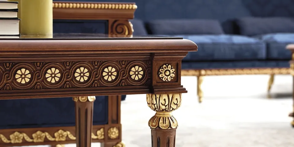 Detalle de una mesa de madera tallada con rosetones dorados junto a un sofá azul